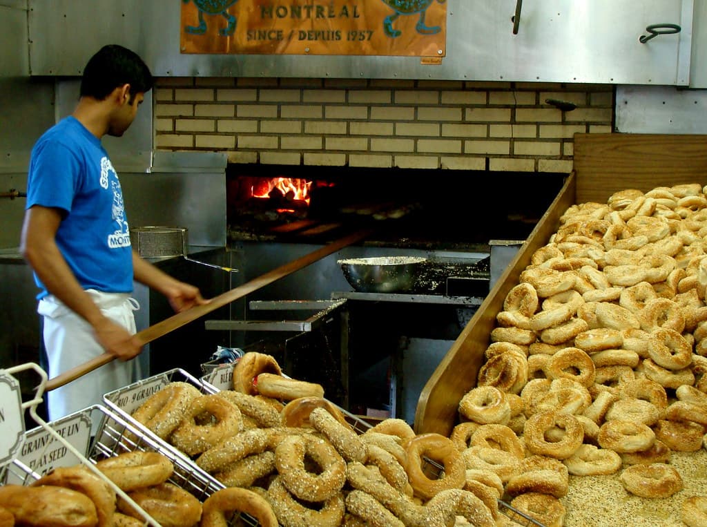 A baker sliding trays of fresh bagels into the oven beside heaped baskets of sesame bagels